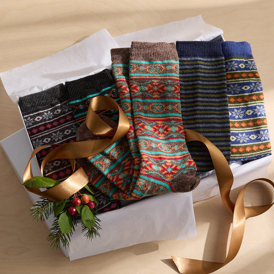 A box of five pairs of patterned socks with festive designs, a gold ribbon, and a small sprig of greenery with red berries on a light wooden surface.