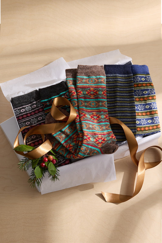 A box of five pairs of patterned socks with festive designs, a gold ribbon, and a small sprig of greenery with red berries on a light wooden surface.