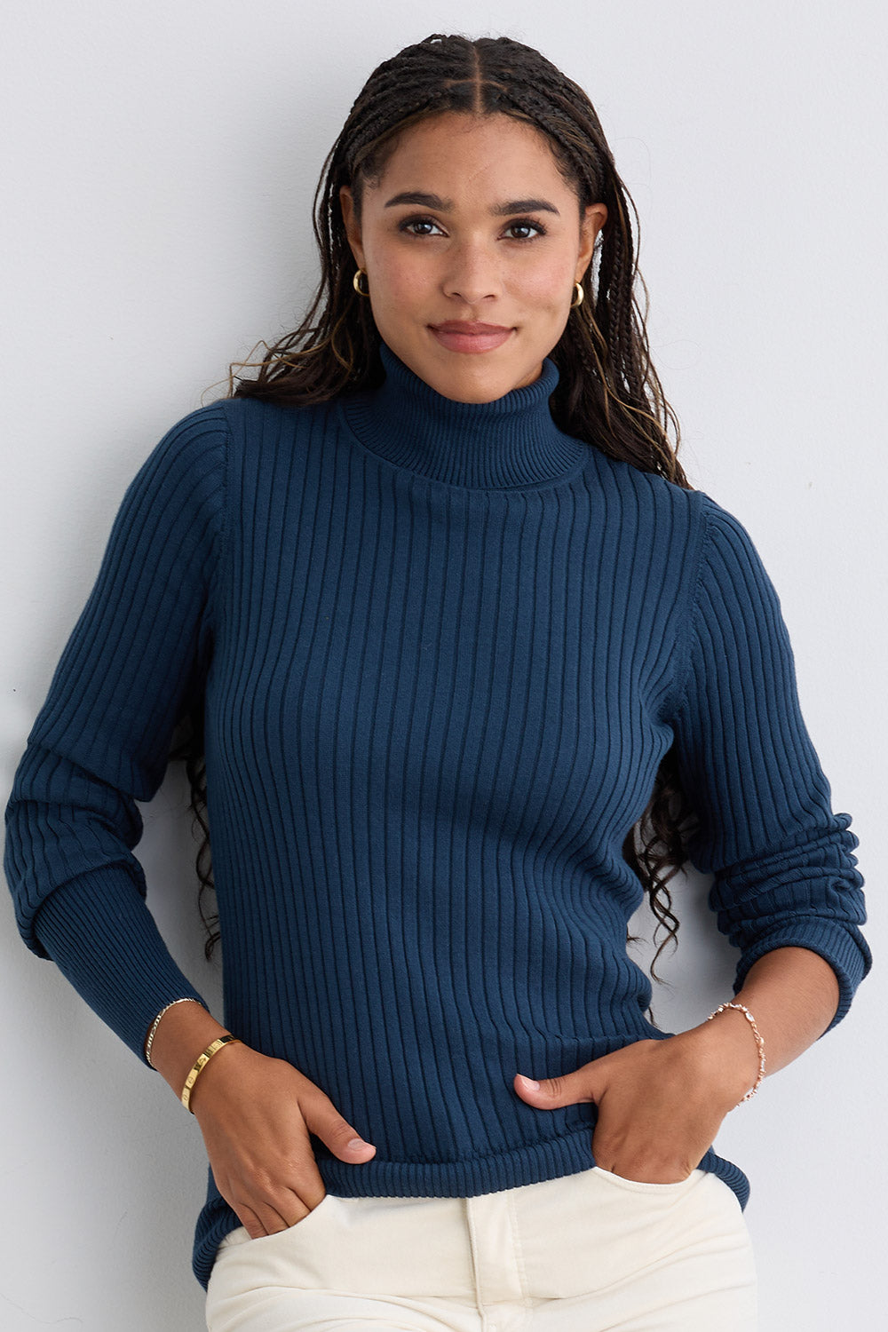 A woman with long braided hair wears a blue ribbed turtleneck sweater and cream pants, standing against a light-colored wall with her hands in her pockets.
