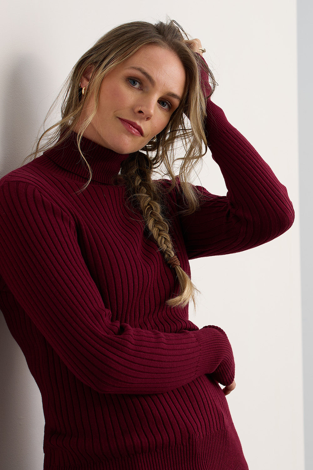 Woman with long, braided hair wearing a ribbed, burgundy turtleneck sweater, standing against a white wall and looking at the camera.