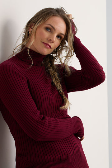 Woman with long, braided hair wearing a ribbed, burgundy turtleneck sweater, standing against a white wall and looking at the camera.