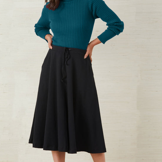 Woman standing against a plain background, wearing a teal ribbed turtleneck sweater, a black A-line midi skirt, and beige heeled sandals.