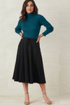 Woman standing against a plain background, wearing a teal ribbed turtleneck sweater, a black A-line midi skirt, and beige heeled sandals.