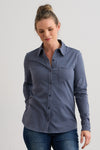 A woman wearing a long-sleeve, button-up, blue shirt with a chest pocket and blue jeans, standing against a plain light-colored background.