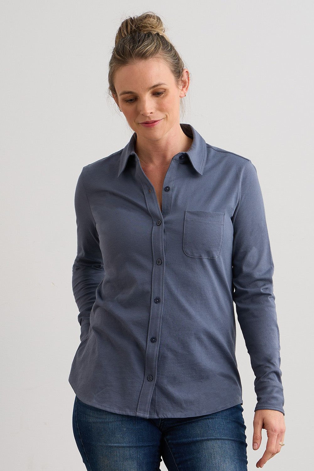 A woman wearing a long-sleeve, button-up, blue shirt with a chest pocket and blue jeans, standing against a plain light-colored background.