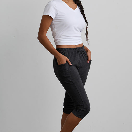 A woman stands barefoot indoors, wearing a white V-neck T-shirt and dark gray cropped jogger pants with her hands in the pockets, facing sideways.