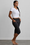 A woman stands barefoot indoors, wearing a white V-neck T-shirt and dark gray cropped jogger pants with her hands in the pockets, facing sideways.