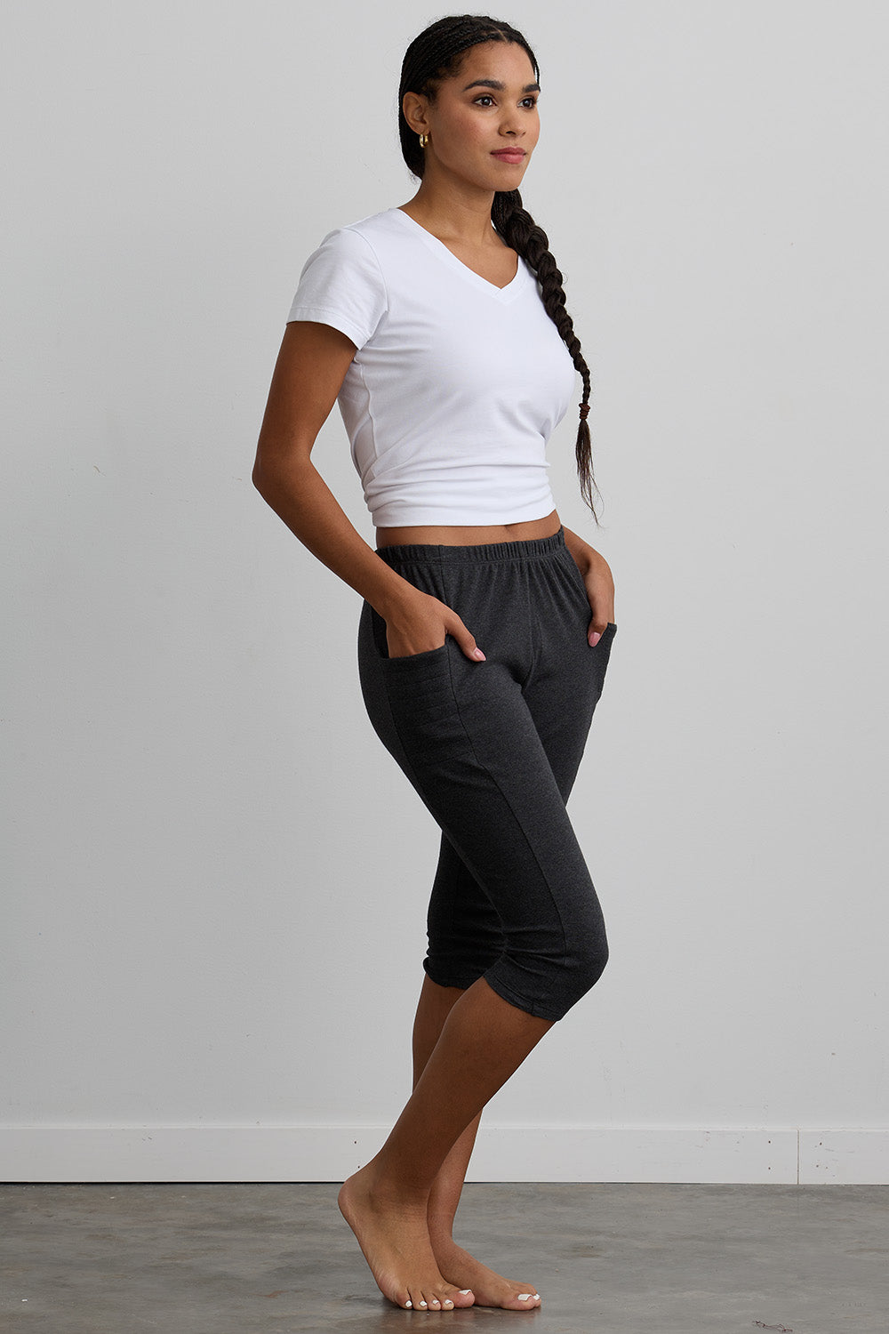 A woman stands barefoot indoors, wearing a white V-neck T-shirt and dark gray cropped jogger pants with her hands in the pockets, facing sideways.