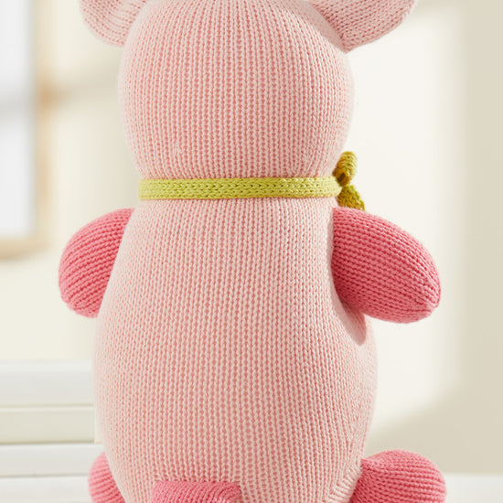 A knitted pink pig plush toy with a green scarf is shown from the back, sitting on a white surface.