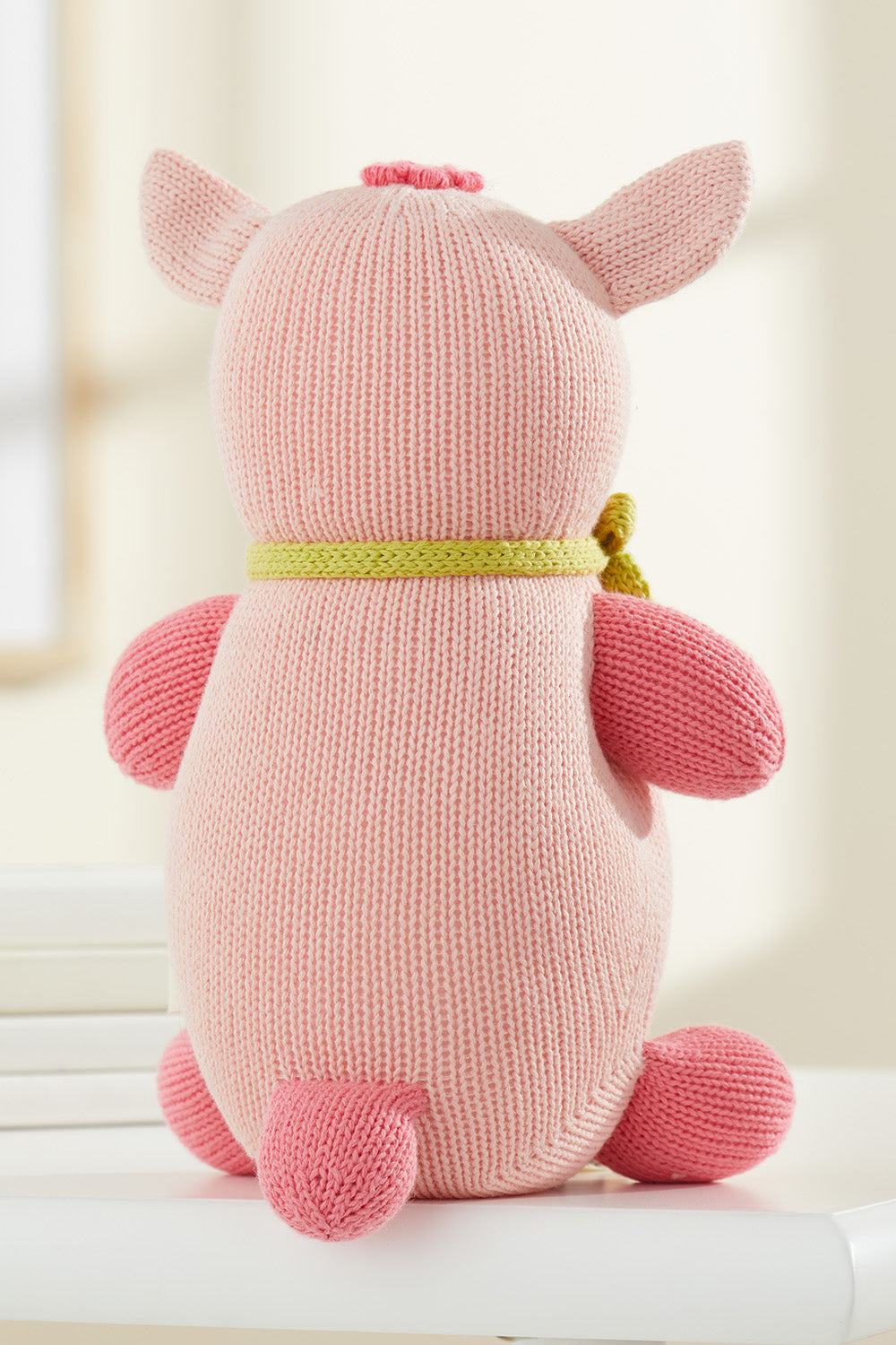 A knitted pink pig plush toy with a green scarf is shown from the back, sitting on a white surface.