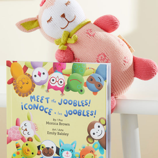 A plush pink and cream stuffed animal leans against a childrens book titled Meet the Joobles! ¬°Conoce a los Joobles! with colorful illustrated animal characters on the cover.