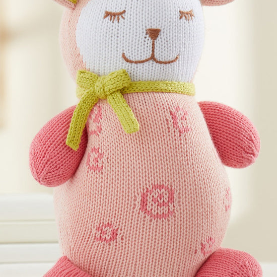 A pink knitted plush toy with closed eyes, a green bow around its neck, and flower patterns on its body sits upright on a white surface.
