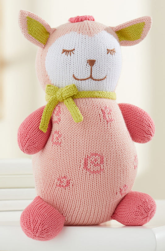 A pink knitted plush toy with closed eyes, a green bow around its neck, and flower patterns on its body sits upright on a white surface.