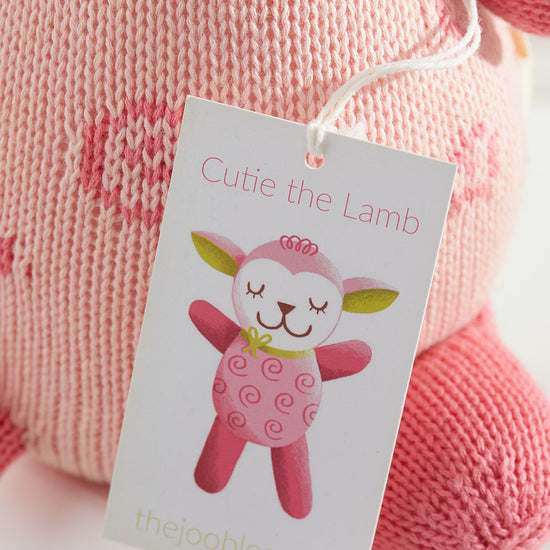 A close-up of a pink knitted stuffed animal with a tag that reads Cutie the Lamb and an illustrated lamb, branded with thejoobles.com.