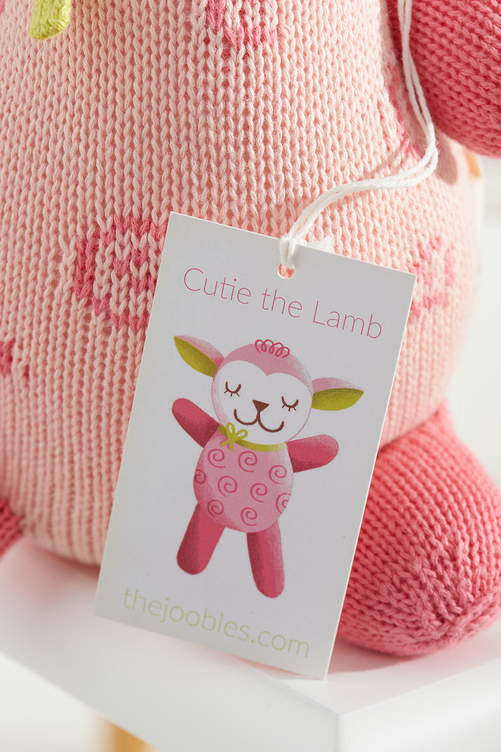 A close-up of a pink knitted stuffed animal with a tag that reads Cutie the Lamb and an illustrated lamb, branded with thejoobles.com.