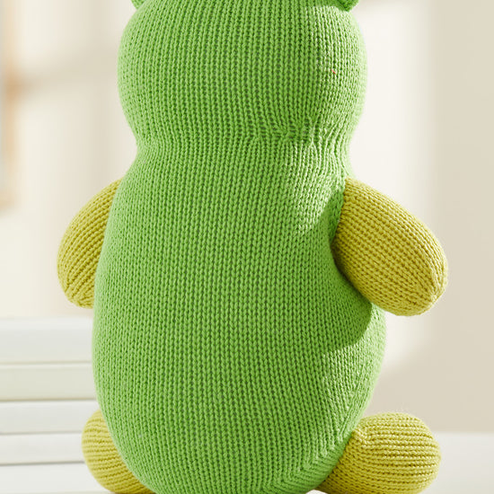 A knitted green and yellow stuffed animal, viewed from the back, sits on a white surface with a neutral background.