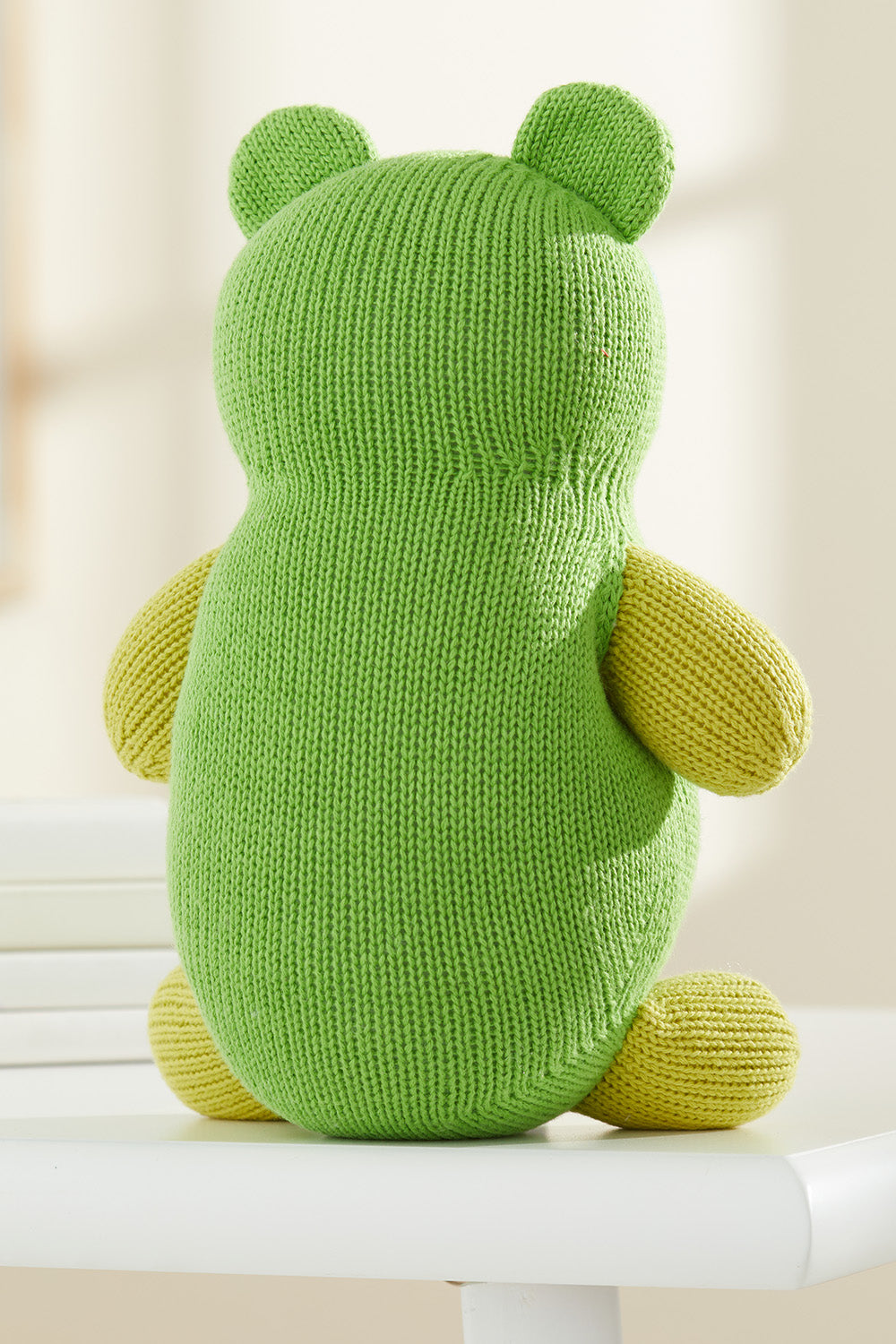 A knitted green and yellow stuffed animal, viewed from the back, sits on a white surface with a neutral background.