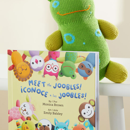A green plush frog sits behind a childrens book titled Meet the Joobles! ¬°Conoce a los Joobles! by Monica Brown, featuring colorful illustrated animal characters on the cover.