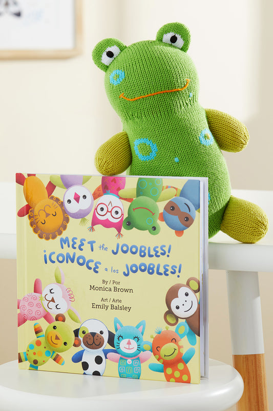 A green plush frog sits behind a childrens book titled Meet the Joobles! ¬°Conoce a los Joobles! by Monica Brown, featuring colorful illustrated animal characters on the cover.
