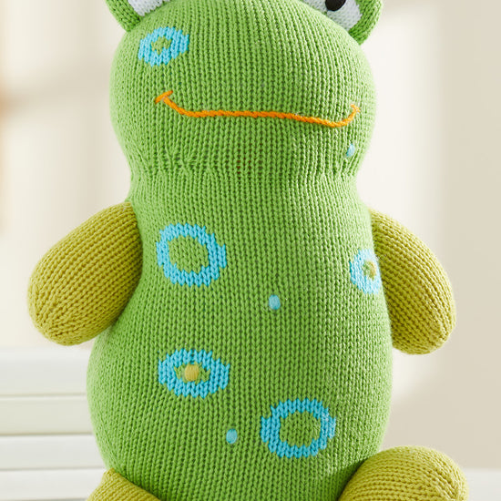 A green knitted frog plush toy with yellow limbs, blue spots, and a simple orange stitched smile sits upright on a white surface.