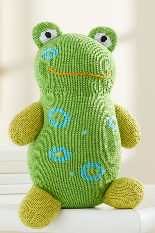 A green knitted frog plush toy with yellow limbs, blue spots, and a simple orange stitched smile sits upright on a white surface.