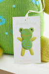 A tag reading Flop the Frog with an illustrated frog is attached to a green knitted toy. The website thejoobles.com is printed at the bottom of the tag.