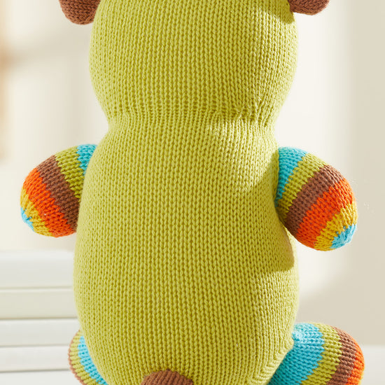 A knitted stuffed animal with a green body, brown ears and tail, and striped arms and legs in blue, green, orange, and brown, shown from the back on a white surface.