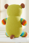 A knitted stuffed animal with a green body, brown ears and tail, and striped arms and legs in blue, green, orange, and brown, shown from the back on a white surface.