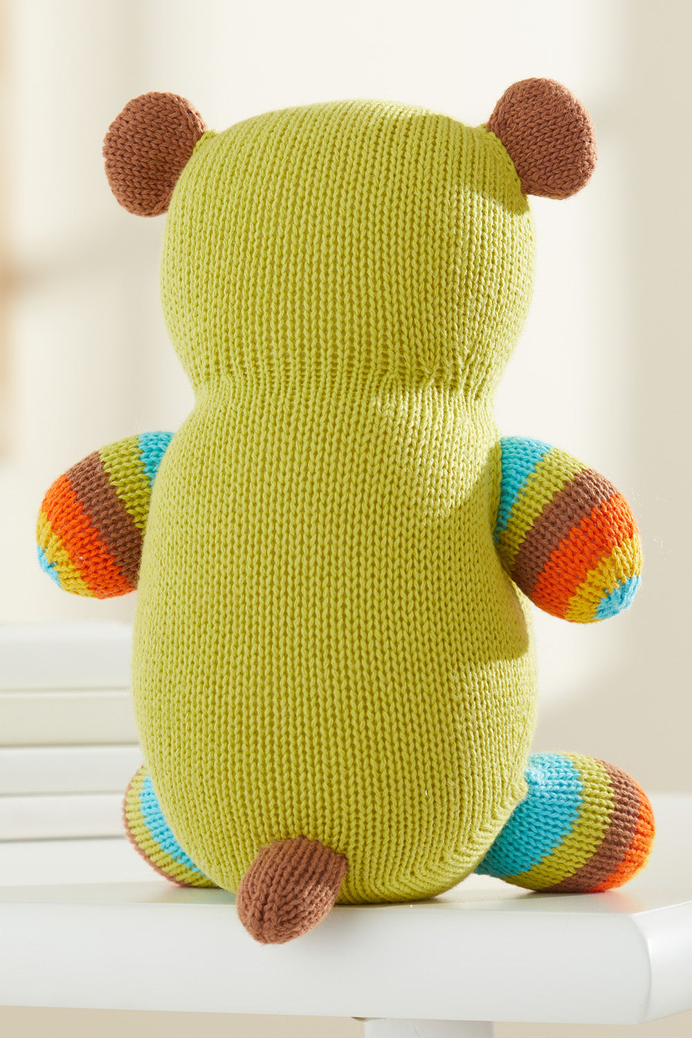 A knitted stuffed animal with a green body, brown ears and tail, and striped arms and legs in blue, green, orange, and brown, shown from the back on a white surface.