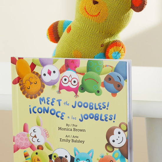 A plush green and orange bear sits behind a childrens book titled Meet the Joobles! ¬°Conoce a los Joobles! featuring colorful animal characters on the cover.