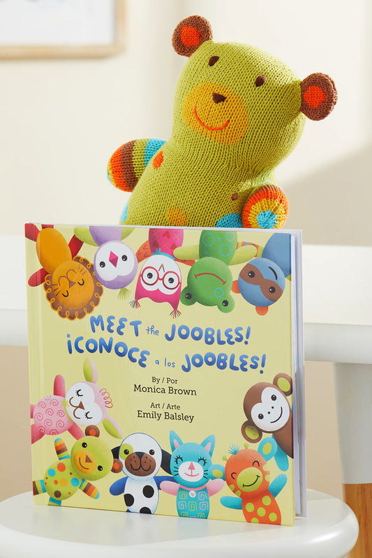 A plush green and orange bear sits behind a childrens book titled Meet the Joobles! ¬°Conoce a los Joobles! featuring colorful animal characters on the cover.