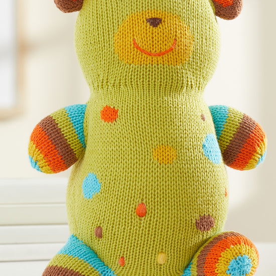 A knitted stuffed bear with green, blue, orange, and brown colors, featuring spots and stripes, sitting on a white surface.