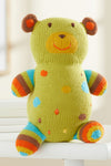 A knitted stuffed bear with green, blue, orange, and brown colors, featuring spots and stripes, sitting on a white surface.