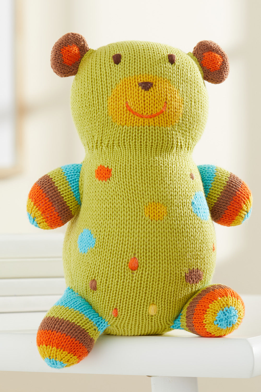 A knitted stuffed bear with green, blue, orange, and brown colors, featuring spots and stripes, sitting on a white surface.