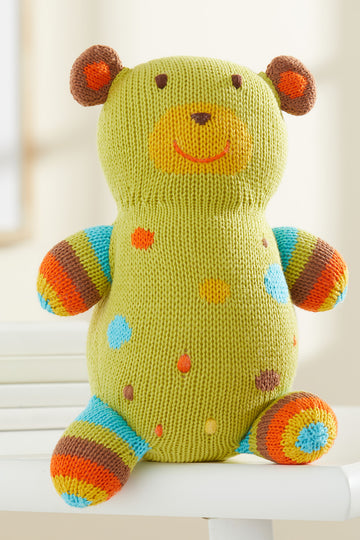 A knitted stuffed bear with green, blue, orange, and brown colors, featuring spots and stripes, sitting on a white surface.