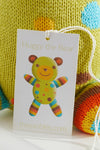 A product tag attached to a knitted toy reads Huggy the Bear with a picture of a colorful bear and the website thejoobles.com below.