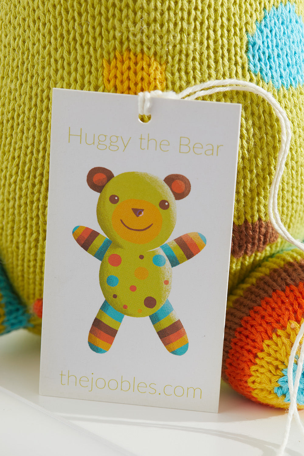 A product tag attached to a knitted toy reads Huggy the Bear with a picture of a colorful bear and the website thejoobles.com below.