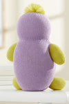 A knitted plush toy with a purple body, yellow arms, legs, and top tuft, sitting on a white surface, facing away from the camera.
