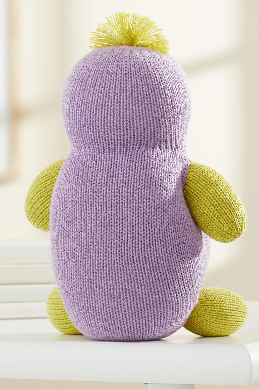 A knitted plush toy with a purple body, yellow arms, legs, and top tuft, sitting on a white surface, facing away from the camera.