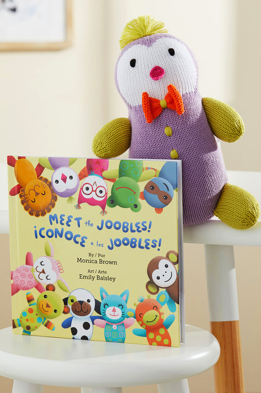 A plush toy in a purple outfit sits on a chair next to a bilingual children‚Äôs book titled Meet the Joobles! ¬°Conoce a los Joobles! featuring colorful animal characters on the cover.