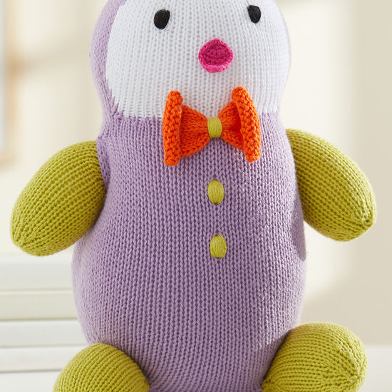 A knitted purple and yellow penguin plush toy with an orange bow tie and two yellow buttons sits on a white surface.