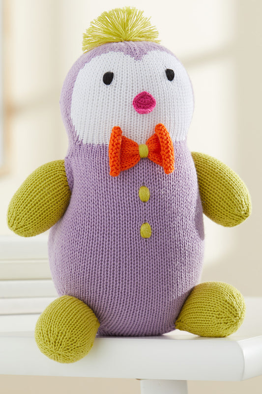 A knitted purple and yellow penguin plush toy with an orange bow tie and two yellow buttons sits on a white surface.