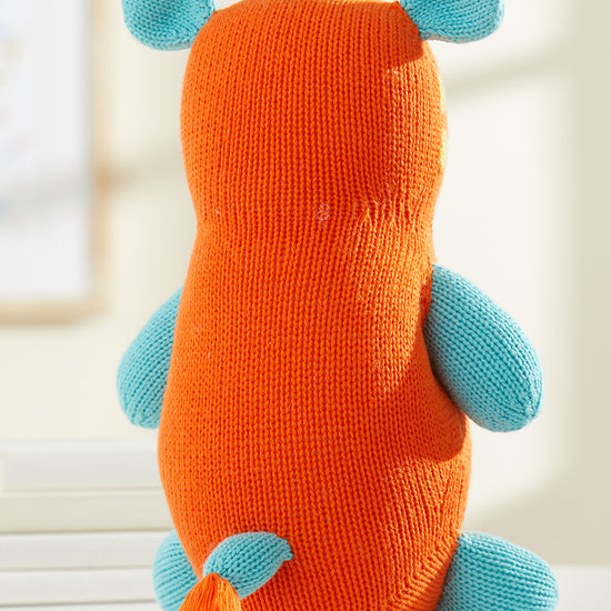 A stuffed toy with an orange body, blue limbs and ears, a blue tail, and a tuft of blue hair sits upright on a white surface.