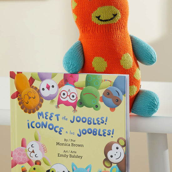 A knit orange and turquoise stuffed animal sits behind a childrens book titled Meet the Joobles! ¬°Conoce a los Joobles! featuring illustrated animal characters on the cover.