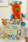 A knit orange and turquoise stuffed animal sits behind a childrens book titled Meet the Joobles! ¬°Conoce a los Joobles! featuring illustrated animal characters on the cover.