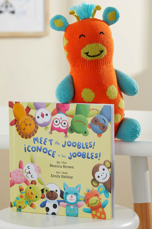 A knit orange and turquoise stuffed animal sits behind a childrens book titled Meet the Joobles! ¬°Conoce a los Joobles! featuring illustrated animal characters on the cover.