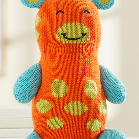 A knitted stuffed toy giraffe with an orange body, blue limbs and ears, yellow spots, a smiling face, and blue yarn hair on top.