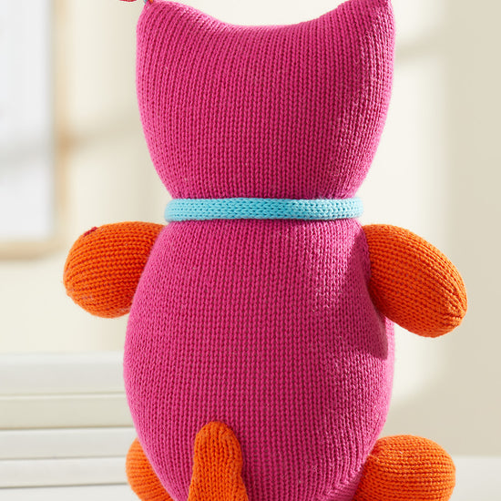 A knitted pink plush toy with orange arms, feet, and a tail, blue collar, and multicolored yarn tassels on its head, sitting on a white surface, seen from behind.