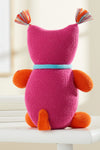A knitted pink plush toy with orange arms, feet, and a tail, blue collar, and multicolored yarn tassels on its head, sitting on a white surface, seen from behind.
