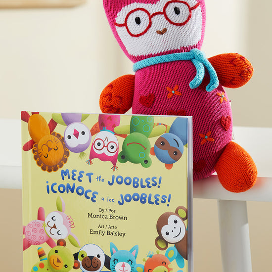 A colorful knit stuffed toy with glasses sits behind a childrens book titled Meet the Joobles! ¬°Conoce a los Joobles! placed on a white chair.